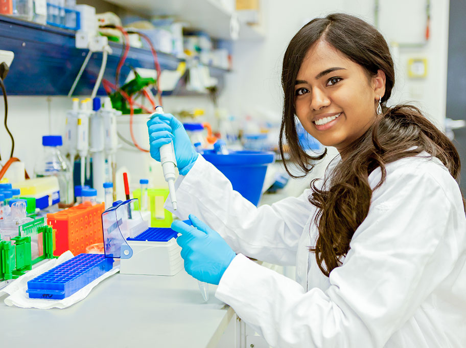 Eine junge Frau mit in weißem Kittel und blauen Gummihandschuhen sitzt im Labor und pipettiert Lösungen.