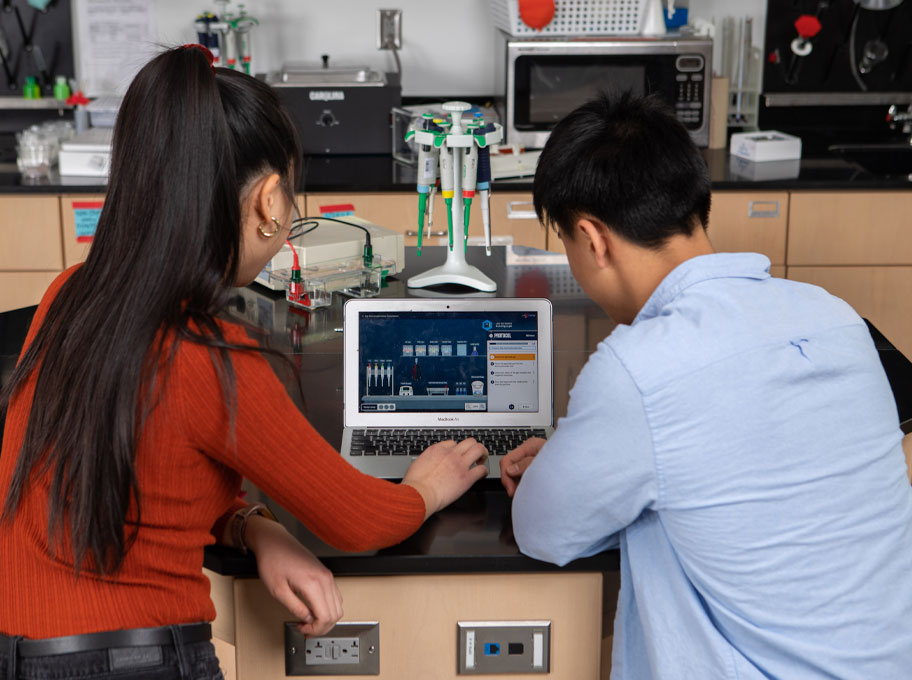 Zwei Jugendliche sitzen im Labor an einem Tisch mit dem Rücken zur Kamera. Sie schauen sich ein Lernvideo auf einem Laptop an.
