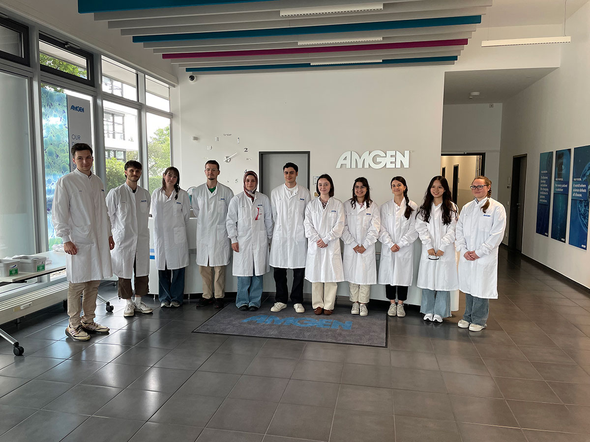 Elf junge Studierende in weißen Kitteln stehen aufgereiht in einem Eingansbereich und lächeln in die Kamera. Im Hintergrund sind das Amgen Logo und einige Plakate zu sehen.