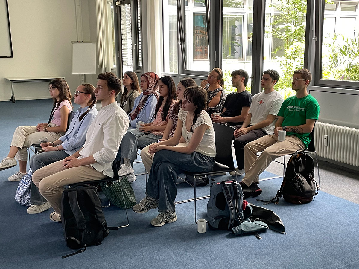 Eine Gruppe von zwölf jungen Studierenden sitzt in drei Reihen auf Stühlen in einem hellen Raum mit großen Fenstern. Die Personen sind aufmerksam nach vorne gerichtet, als würden sie einer Präsentation oder einem Vortrag folgen.