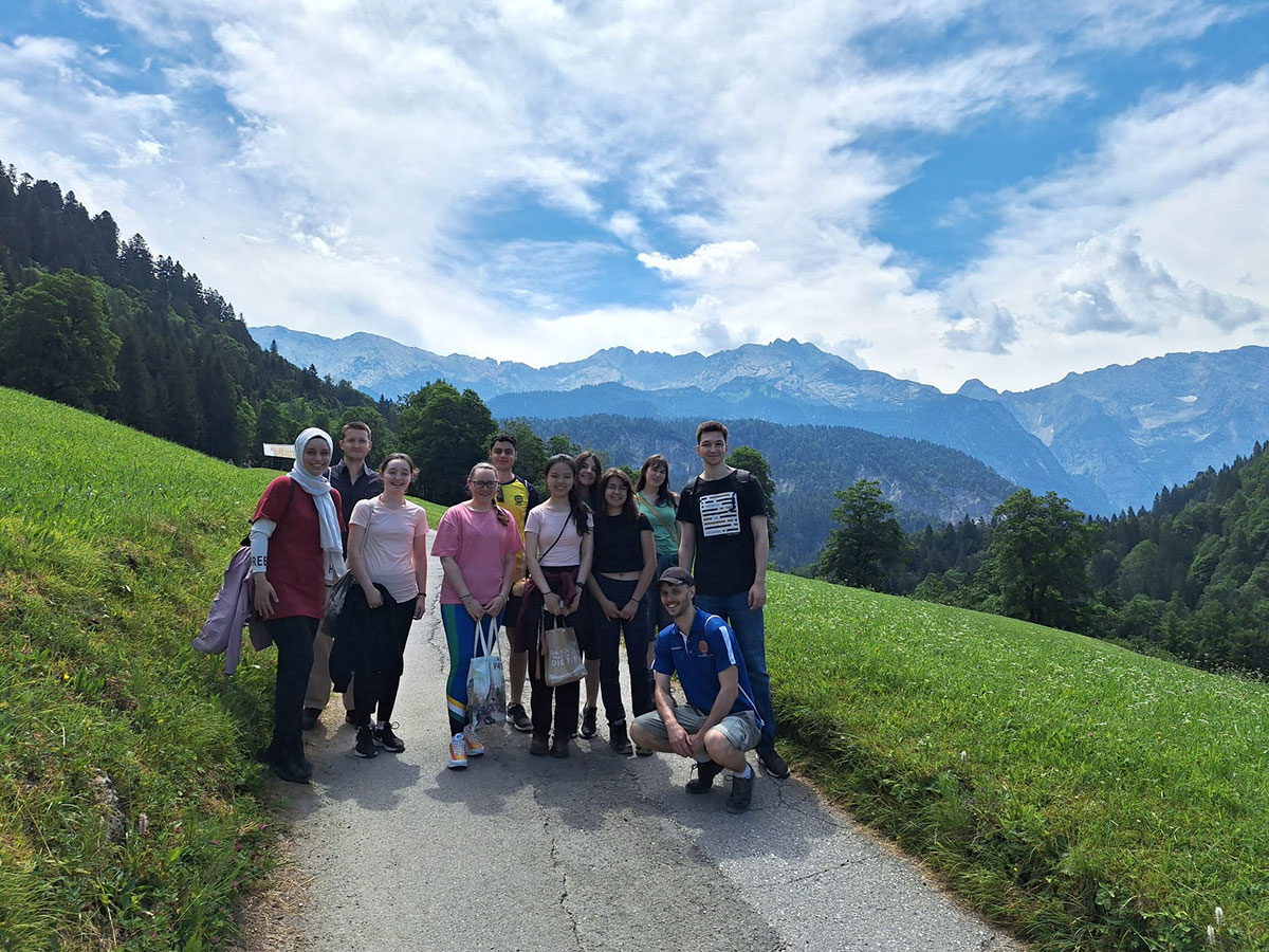Eine Gruppe von elf Studierenden steht auf einem schmalen Weg in einer grünen Berglandschaft mit Wiesen, Wäldern und hohen Bergen im Hintergrund unter blauem Himmel mit Wolken.
