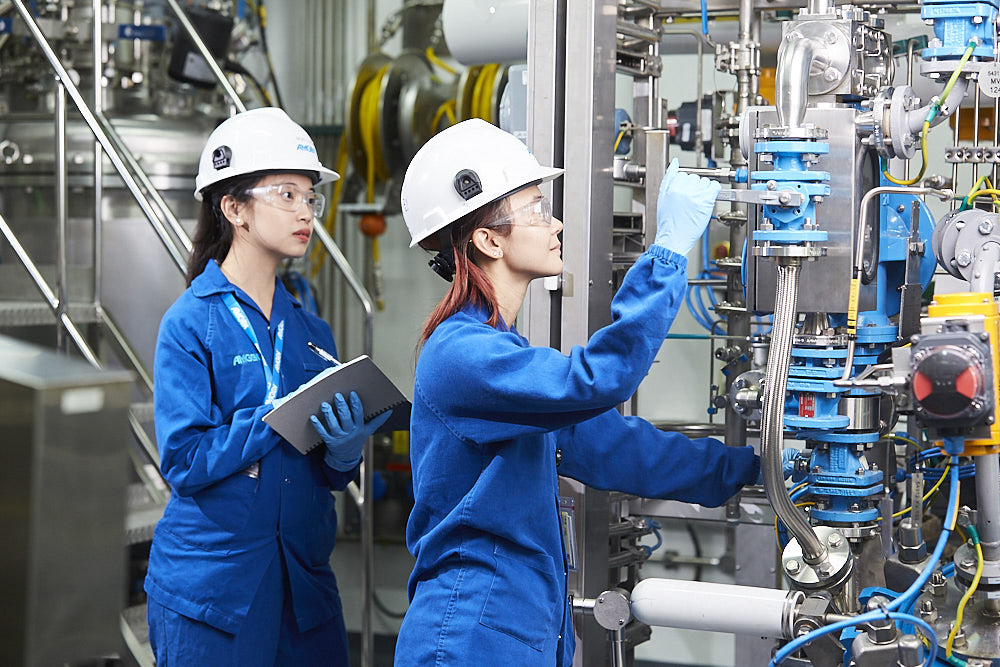 Zwei Mitarbeiterinnen mit blauen Anzügen und weißen Helmen stehen an einer großen Produktionsmaschine. Während eine Frau an der Maschine arbeitet, notiert die andere Informationen auf einem Schreibblock.